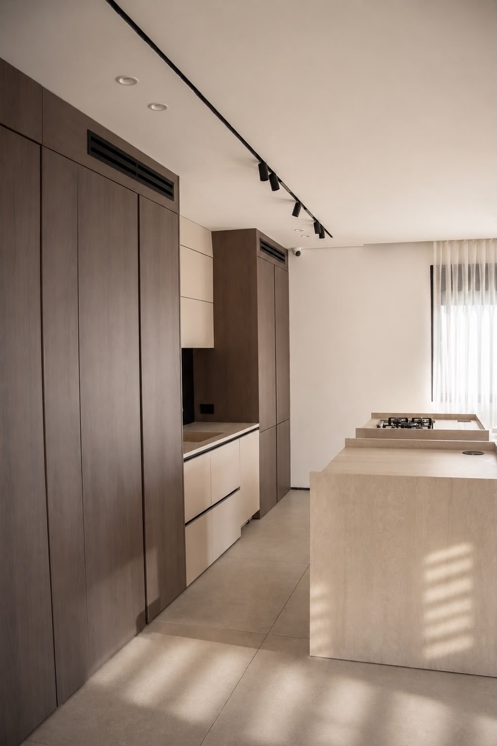 A completed kitchen: full-height dark-oak cabinetry with vent grilles, warm-toned lower cabinets, a travertine-toned island and track lighting.