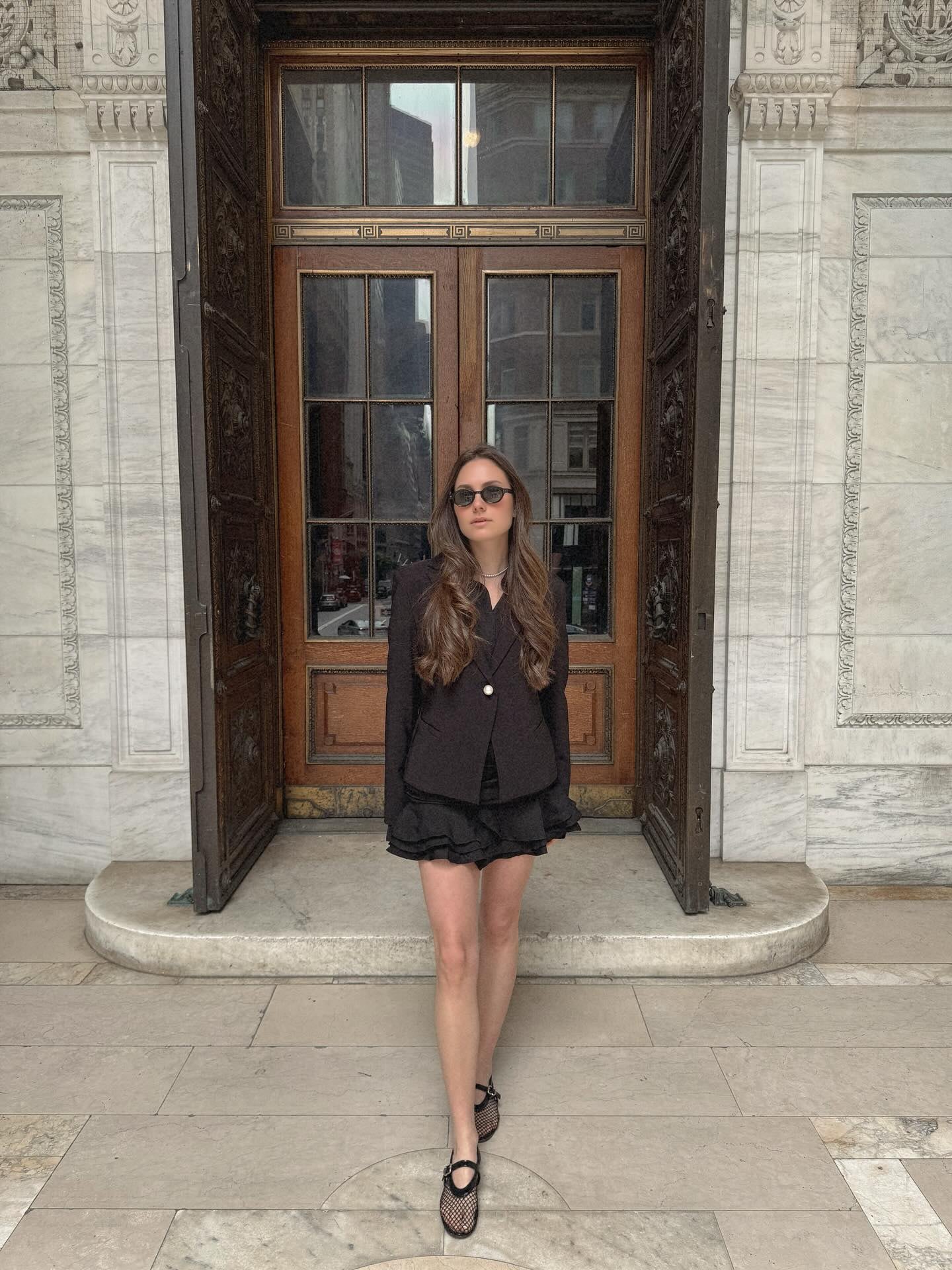 Shoham Sela at a monumental doorway in New York: open carved-bronze doors, wood-and-glass inner doors with a brass band, white marble walls and a rounded stone step.